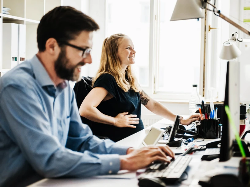 Eine schwangere Frau und ihr Arbeitskollege sitzen im Büro an ihrem Arbeitsplatz