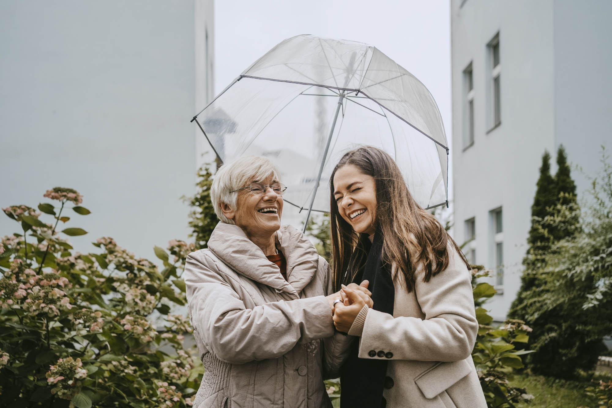 Rentnerin mit junger Frau lachend unter einem Schirm im Vorhof von Wohnblöcken<br/>