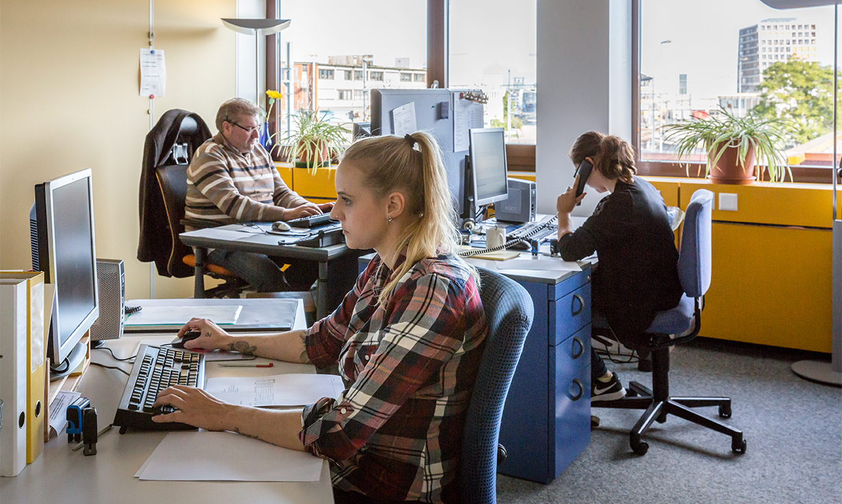 Ein Büro mit drei Schreibtischen, an denen Personen arbeiten.